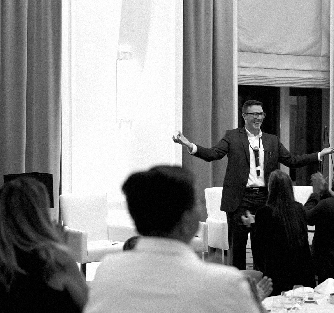 Black-and-white photo from the back of a banquet room: guests at dinner tables watch a performer at the front of the room at the front of the room.