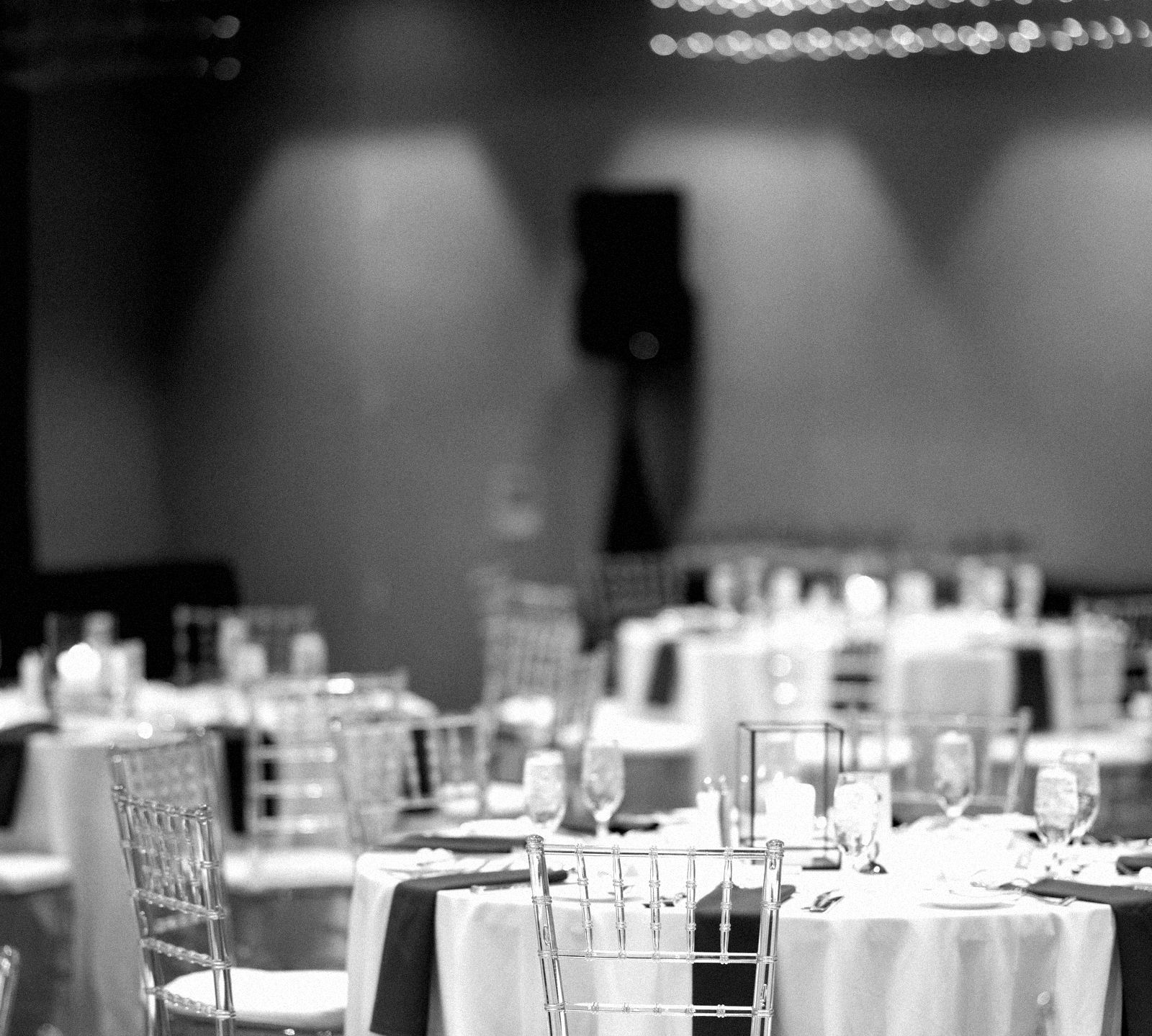 An empty hotel ballroom before a corporate event: round tables set with white linens, navy napkins and candles under strings of ceiling lights.