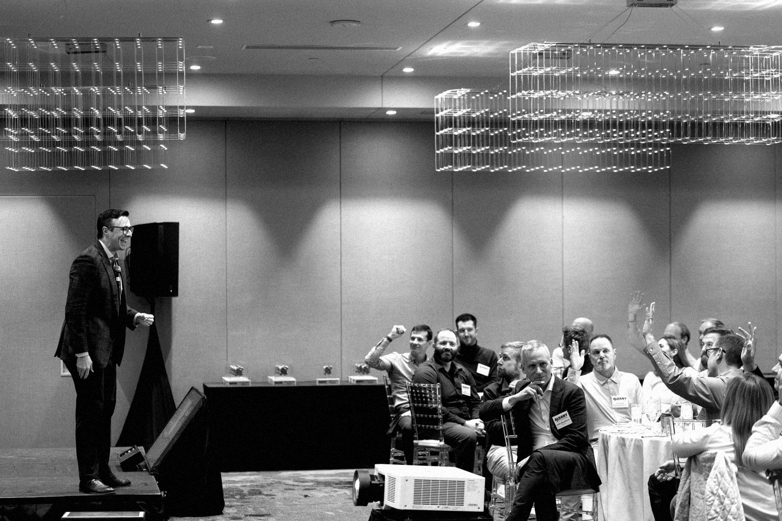 Black-and-white photo of a corporate audience at round tables raising their hands as a performer in a blazer laughs from a low stage beside a speaker.