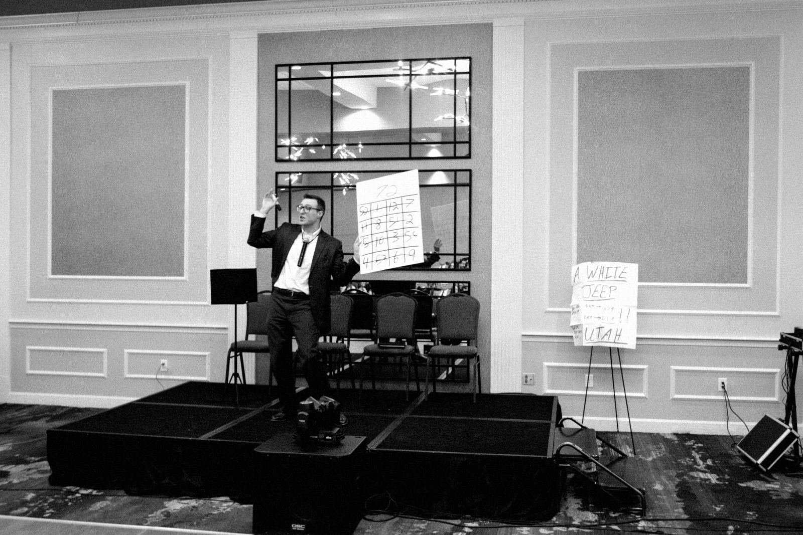 Wide shot of a small black stage in a hotel function room, a performer holding up a large handwritten number grid while a few guests watch from chairs.